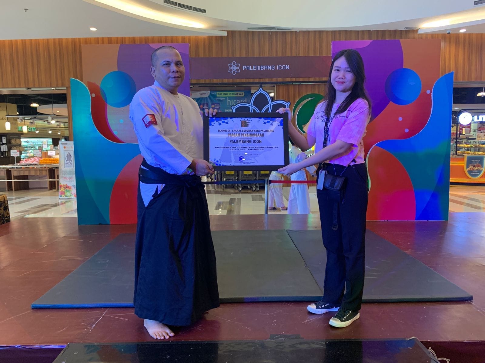 Ni Made Rianita Hariyanti leading an Aikido event at Palembang Icon Mall, presenting a certificate on stage during the ceremony.